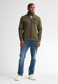 Olijfgroene jas met hoge kraag, frontale rits en textielstructuur. Voorzien van een borstzak; gecombineerd met lichtblauwe jeans en witte sneakers.
