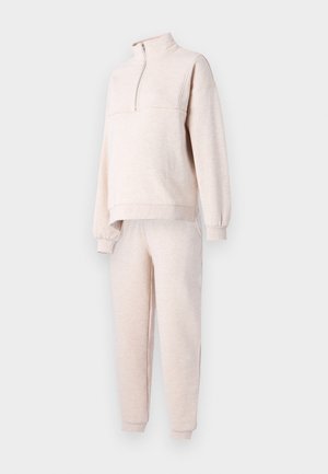 Jasno beżowy dres z polaru, z wysokim kołnierzem na zamek, luźnym krojem, ściągaczami przy mankietach i w pasie oraz dopasowanymi spodniami.