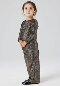 Sprankelend bruin stoffen tweedelig outfit met lange mouwen en loszittende broeken, gecombineerd met zwarte schoenen. Over het geheel een gladde textuur.