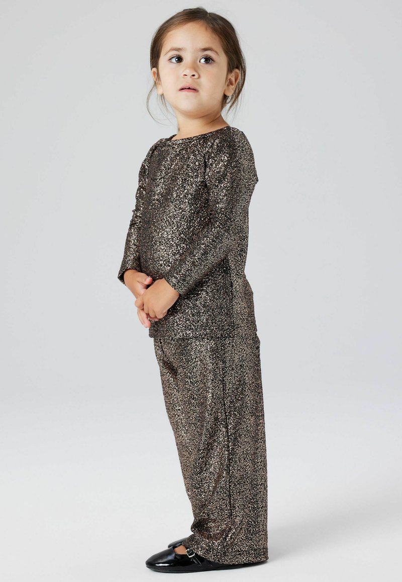 Sprankelend bruin stoffen tweedelig outfit met lange mouwen en loszittende broeken, gecombineerd met zwarte schoenen. Over het geheel een gladde textuur.