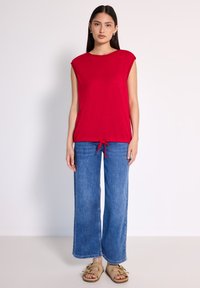 Top sin mangas rojo con escote redondo y dobladillo con cordón, combinado con jeans de denim azul de pierna ancha y sandalias beige.