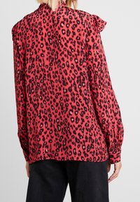 Red blouse with black leopard print, ruffled shoulders, and long sleeves. Made of smooth material, featuring a slightly loose fit and gathered cuffs.
