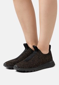 Zwarte instappersneakers met een gestructureerde geweven bovenkant met metallic gouden spikkels, elastische accenten en zwarte rubberen zolen voor grip.