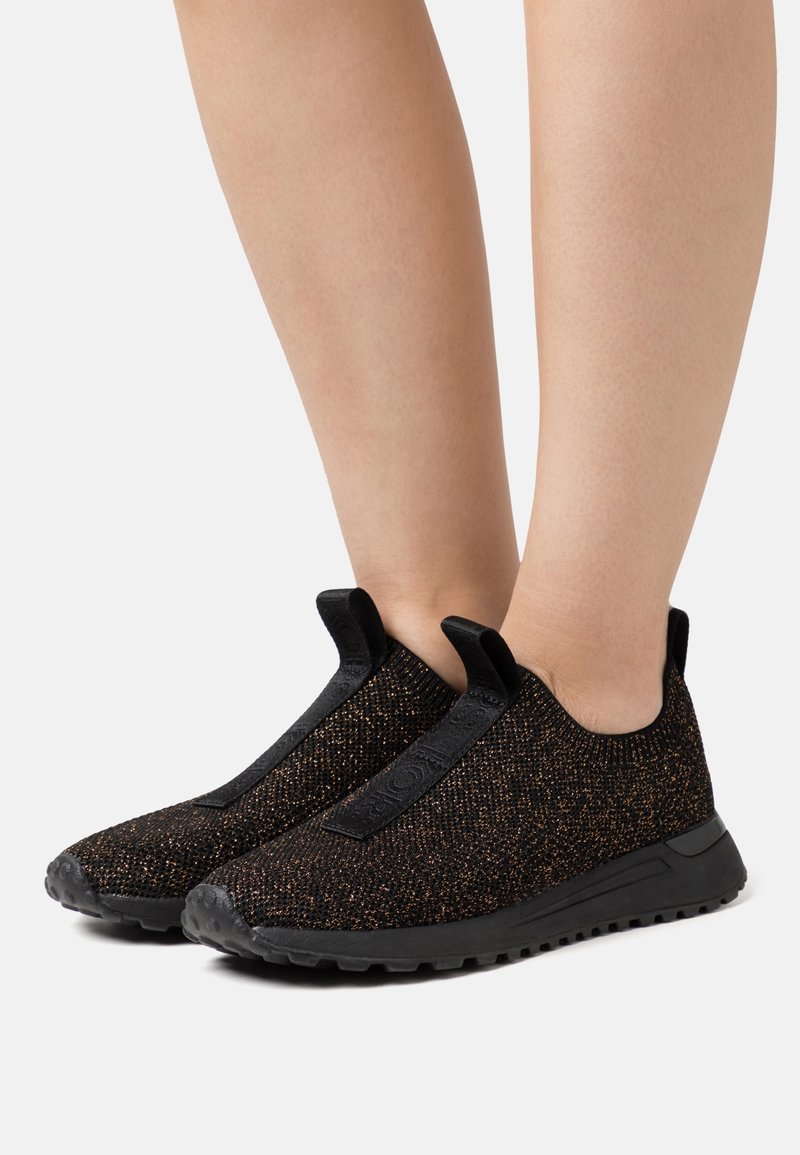 Zwarte instappersneakers met een gestructureerde geweven bovenkant met metallic gouden spikkels, elastische accenten en zwarte rubberen zolen voor grip.