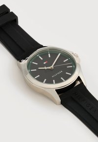 Ασημένιο ρολόι χειρός με μαύρο καντράν, λευκούς δείκτες ώρας και μαύρο λουράκι από σιλικόνη με το εμπορικό σήμα "Hilfiger".