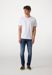 White cotton t-shirt with a small triangular logo on the chest, paired with blue jeans and brown suede shoes. Model poses standing.