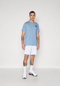 Tricou scurt de culoare albastru deschis, cu un mic logo brodat. Asortat cu pantaloni scurți albi și adidași albi cu accente negre și roșii.