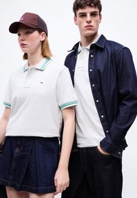 Νεαρή γυναίκα με λευκό πόλο με πράσινες ρίγες και καφέ καπέλο στέκεται δίπλα σε νεαρό άνδρα με λευκό πόλο και σκούρο τζιν μπουφάν και παντελόνι.