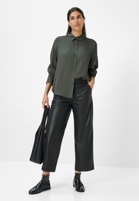 Donna in camicia verde scuro con bottoni, pantaloni in pelle larghi neri, mocassini neri e calzini luccicanti, mentre tiene una borsa tote nera.