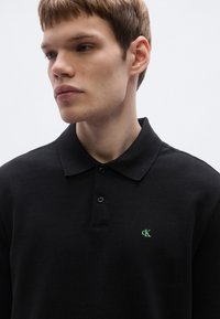 Mustast tekstuuriga kangast polo särk, millel on vasakul rinnal roheline tikitud logo ja klassikaline krae kahe nupuga.