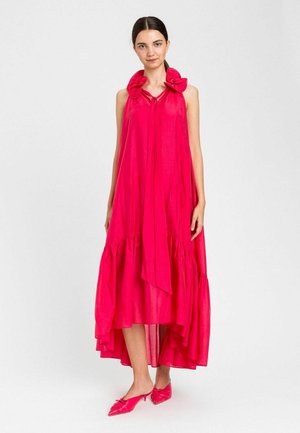 Donna che indossa un vestito senza maniche rosa acceso con orlo più lungo dietro, decorato con grandi motivi floreali sulle spalle, e décolleté rosa con tacco a spillo basso e punta appuntita abbinate.