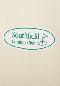 Kremowa tkanina z owalną białą metką z zielonym tekstem: "Southfield Country Club" oraz emblematem w postaci flagi golfowej.