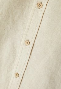 Chemise en lin beige avec une texture lisse, dotée de plusieurs boutons le long de la patte de boutonnage dans une finition en bois clair et de subtils détails de couture.