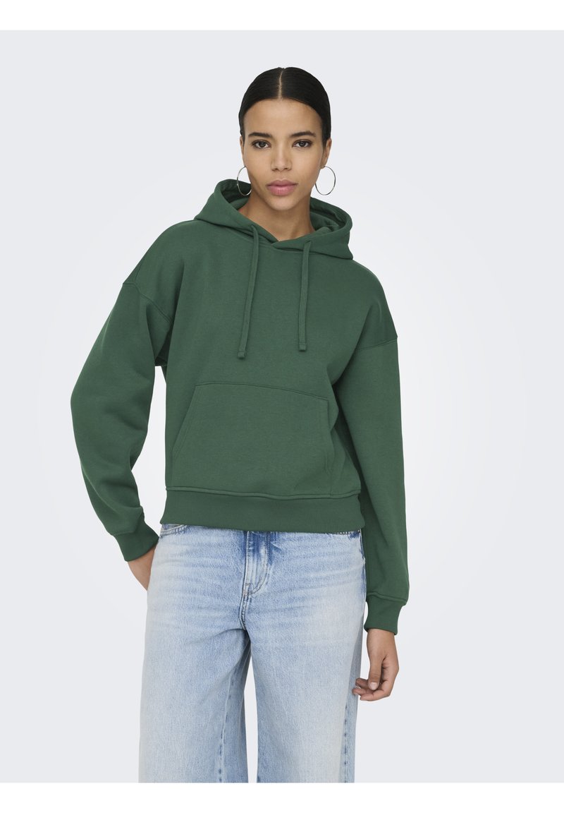 Femme aux cheveux foncés portant un sweat à capuche vert et un jean bleu clair, debout devant un fond clair uni, regardant droit devant elle.