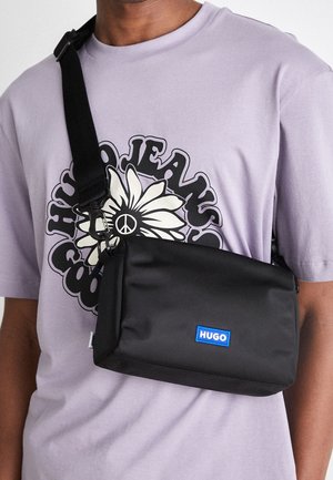 Mann trägt ein hellviolettes T-Shirt mit weißem Blumen- und Friedenszeichen-Grafik, trägt eine schwarze Hugo Crossbody-Tasche mit verstellbarem Riemen.