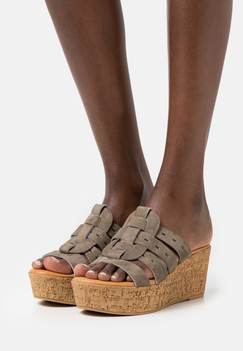 Pieds portant des sandales compensées taupe en daim avec brides tissées et semelles compensées en liège sur fond blanc.