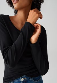 Maglietta nera a maniche lunghe con colletto a V, realizzata in tessuto liscio. Presenta maniche aderenti e una leggera elasticità, abbinata a jeans blu.