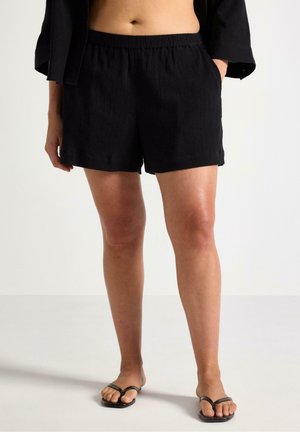 Person iført sorte teksturerede shorts, sort åben-front top og sorte thongsandaler, stående mod en ensfarvet lys baggrund.