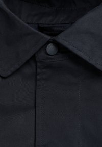 Gros plan sur le col d'une chemise noire avec un bouton noir fermé en haut, montrant les coutures détaillées autour du col et de la patte de boutonnage.