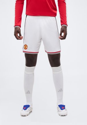 Mężczyzna ubrany w białe spodenki piłkarskie Manchester United, białe podkolanówki, białe korki z niebieskimi nakładkami i czerwoną bluzę z długim rękawem.