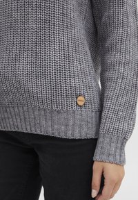 Grau gestrickter Pullover mit strukturiertem Gewebe, gerippten Bündchen und Saum sowie einem Holzmarkenschild an der unteren Seite.
