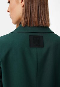 Dark green coat featuring a textured fabric, pointed collar, and a prominent black logo patch on the back. Smooth, tailored design.