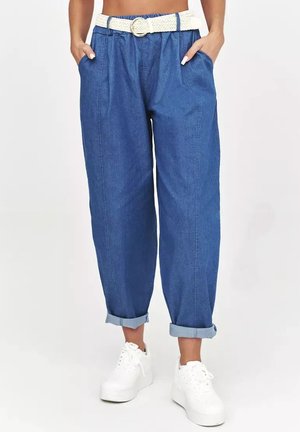 Kobieta nosząca niebieskie dżinsy z wysokim stanem i podwiniętymi nogawkami, biały pleciony pasek, ręce w kieszeniach oraz białe sneakersy na platformie.