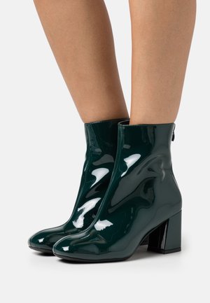 Bottines en cuir verni vert avec un talon carré, texture lisse et détails en zipper, affichant une silhouette élégante et ajustée.