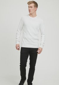 Bela rebrasta pletena pulover s dolgimi rokavi, v kombinaciji s slim fit črnimi kavbojkami in črnimi supergami, kar ustvarja preprost, sodoben videz.