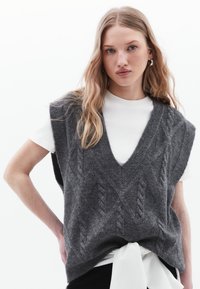 Gilet grigio a maglia con scollatura a V, maniche corte e motivi a treccia. Abbinato a una camicia bianca con un dettaglio legato in vita.