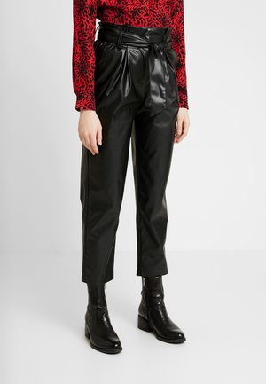 Femme portant un pantalon noir en similicuir taille haute avec une ceinture nouée, associé à des bottines noires et un haut à manches longues rouge à motif.