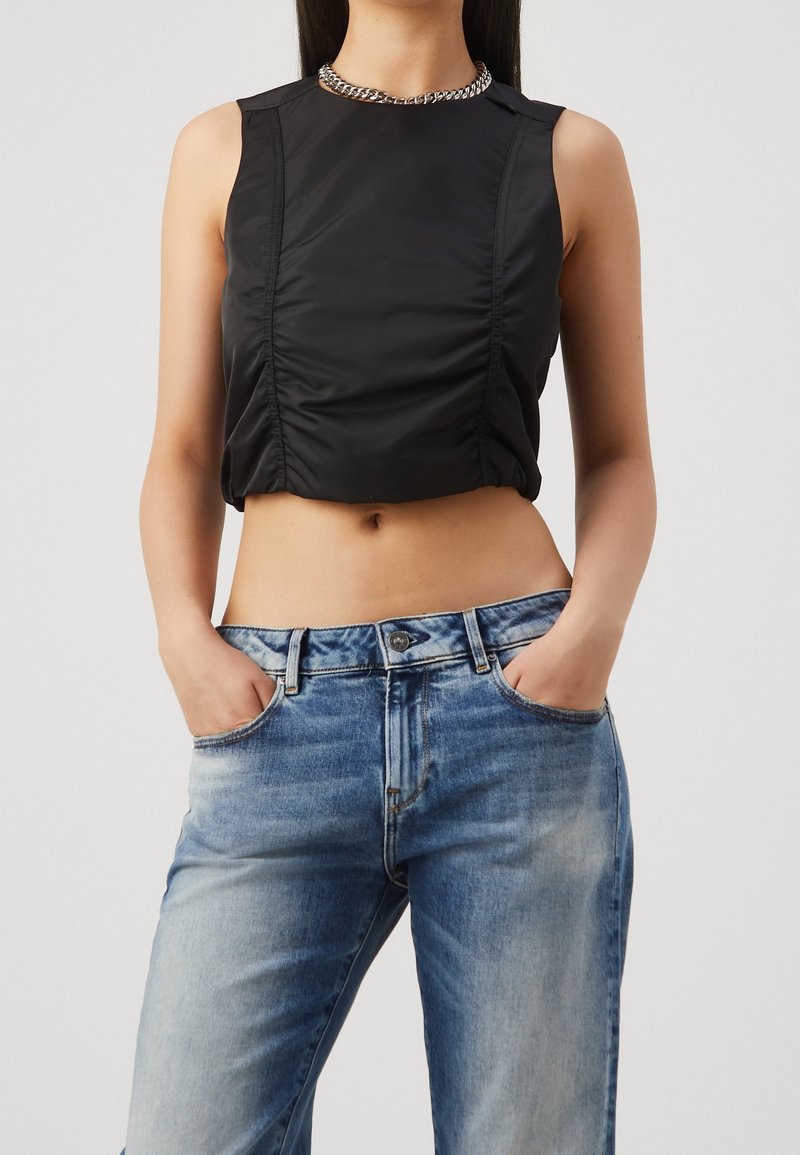 Femme portant un top court noir sans manches avec des coutures froncées et un jean délavé bleu clair, les mains dans les poches.