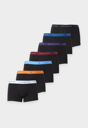 Homme debout torse nu, bras croisés, portant un boxer noir avec une ceinture blanche et bleue Tommy Hilfiger sur un fond uni.