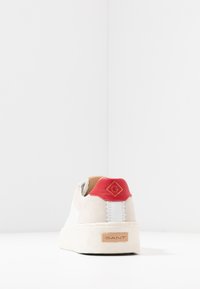 White suede sneaker with a red leather heel accent, brown logo patch, and light-colored rubber sole. Features white side stripes.