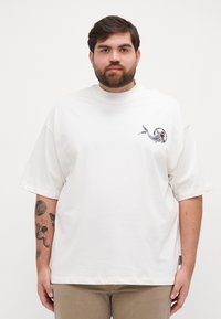 White oversized t-shirt with short sleeves, featuring a colorful fish graphic on the left chest area. Smooth texture and casual fit.