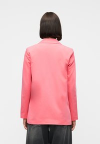 Personne aux cheveux courts et foncés portant un blazer rose à manches longues et un pantalon gris foncé, tournée de dos sur un fond clair uni.