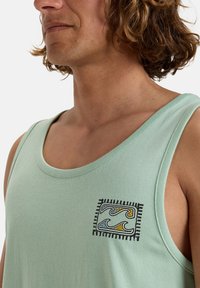 Hellgrünes Tanktop aus weicher Baumwolle, mit einem quadratischen, gestickten Design von Wellen in Blau, Gelb und Schwarz.