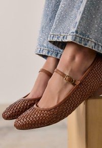 Mocasines de cuero trenzado marrón con punta redondeada, tira para el tobillo y pequeño hebilla dorada, combinados con unos vaqueros de mezclilla azul claros con un dobladillo deshilachado.