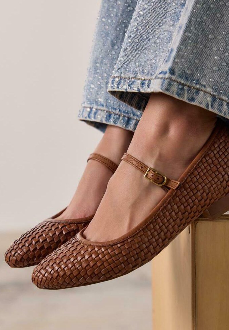 Mocasines de cuero trenzado marrón con punta redondeada, tira para el tobillo y pequeño hebilla dorada, combinados con unos vaqueros de mezclilla azul claros con un dobladillo deshilachado.