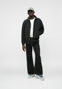 Melna bomber jaka, balts T-krekls, tumšas bikses, bežs cepure un sporta apavi ar zilām akcentiem. Galvenās iezīmes ietver brīvu piegriezumu un monohromatisku krāsu paleti.