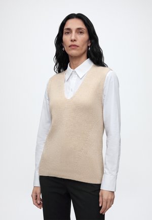 Vrouw met lang donker haar die zilveren creolen draagt, een witte blouse met kraag, een beige gebreid mouwloos vest en een zwarte broek tegen een effen achtergrond.