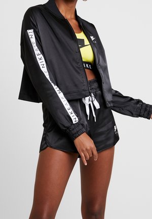 Zwarte Nike cropped jas met witte logo streep op de mouwen, gecombineerd met zwarte Nike shorts en een neon gele Nike sportbeha.