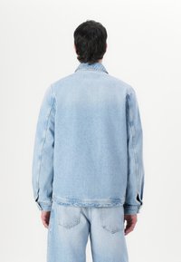 Lyseblå denimjakke med rett snitt, knapper på ermene og klassisk krage. Har en lett teksturert overflate og synlige sømmer.