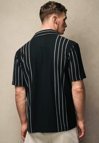 Camisa negra con textura acanalada que presenta rayas verticales blancas y un cuello tipo solapa. Se aprecian mangas cortas y un corte relajado.