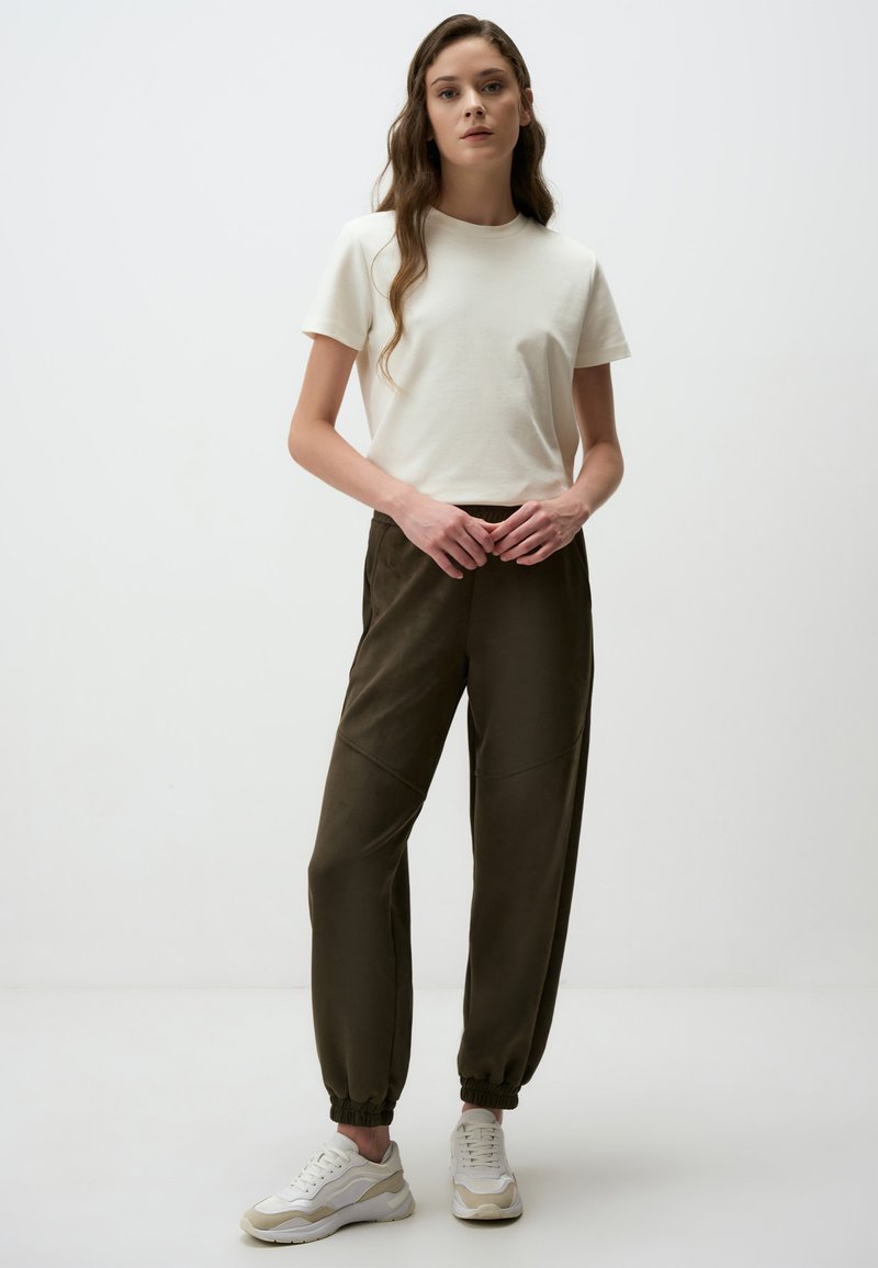 Weißes Baumwoll-T-Shirt kombiniert mit dunkelgrünen Jogginghosen mit elastischen Bündchen und seitlichen Details. Die Schuhe sind weiße Sneaker mit beige Akzenten.