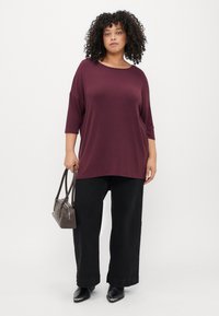 Blusa larga em bordeaux com mangas três quartos, combinada com calças pretas de perna larga e uma pequena bolsa cinza. Sapatos pretos de bico fino.