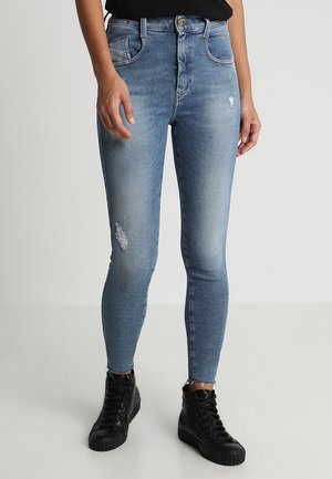 Person trägt eng anliegende, hellblaue, verwaschene Jeans und schwarze High-Top-Sneaker, steht vor einem einfarbigen hellgrauen Hintergrund.