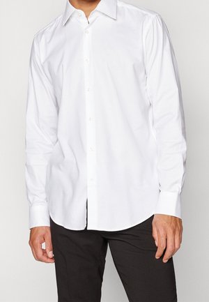 Homme portant une chemise blanche à manches longues boutonnée et un pantalon noir, visible des épaules aux hanches sur un fond uni.
