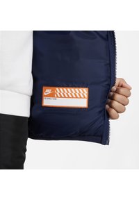Interno di giacca blu navy con un'etichetta arancione che riporta "NOME PREFERITO" e accenti a righe. Chiusura con zip e tessuto strutturato.