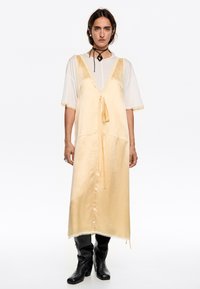 Abito giallo di satin con scollatura a V profonda e fiocco frontale, sovrapposto a una camicia bianca a maniche corte. Stivali neri al ginocchio completano il look.